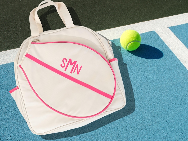 May include: A white tennis bag with a pink trim and the initials "SMN" in pink lettering. The bag is shaped like a tennis racket and is lying on a blue tennis court with a yellow tennis ball.