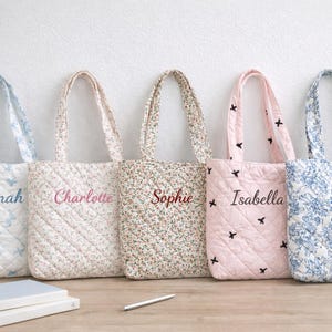 May include: Five quilted tote bags with floral and patterned designs. Each bag has a different colour scheme and a name embroidered on the front: Hannah, Charlotte, Sophie, Isabella, and Mia. The bags are on a wooden surface.