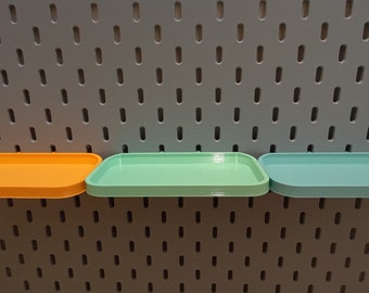 Könnte beinhalten: Drei rechteckige Tabletts in Orange, Hellgrün und Türkisblau sind an einer grauen Lochwand montiert.
