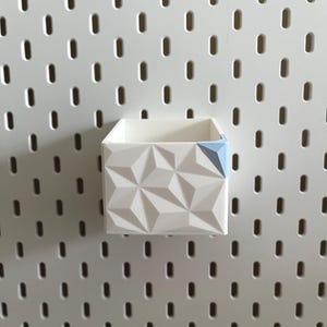 Puede incluir: Un contenedor de almacenamiento geométrico blanco con un acento azul, montado en una tabla perforada blanca. El contenedor tiene un diseño texturizado y facetado. La tabla perforada tiene agujeros ovalados espaciados.