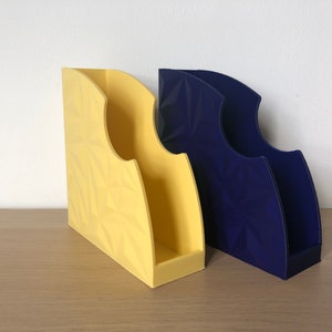 May include: Two magazine holders, one yellow and one blue, with a geometric pattern. The holders are made of plastic and have a modern design.