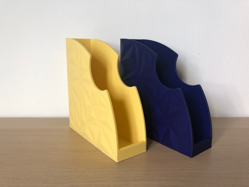 May include: Two magazine holders, one yellow and one blue, with a geometric pattern. The holders are made of plastic and have a modern design.