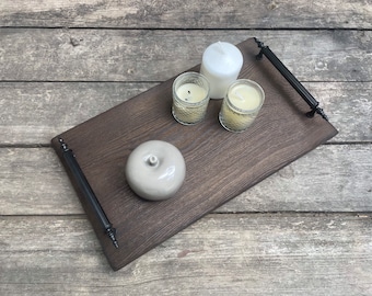 May include: A rectangular dark brown wooden serving tray featuring two black metal handles on opposite ends. The tray holds a smooth light gray ceramic apple-shaped object, two small glass votive candles, and one larger white pillar candle.