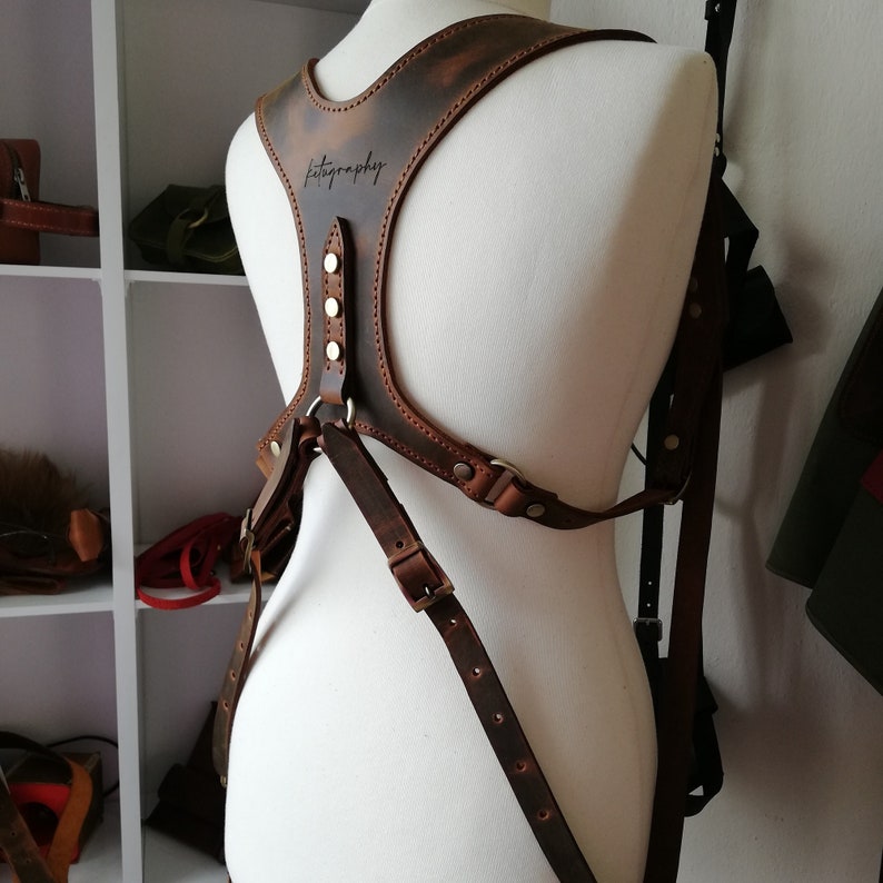 May include: A brown leather camera harness with the word "fotography" inscribed on the chest piece. The harness features adjustable straps, metal hardware, and a distressed finish. The harness is displayed on a mannequin.