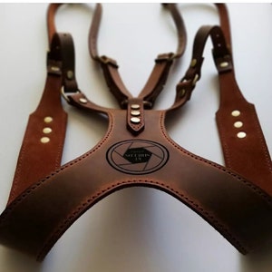 May include: A brown leather camera harness with adjustable straps and brass rivets. The harness features a central chest piece with an oval logo that reads "KL Shields Photography". The design includes multiple connection points for attaching cameras.
