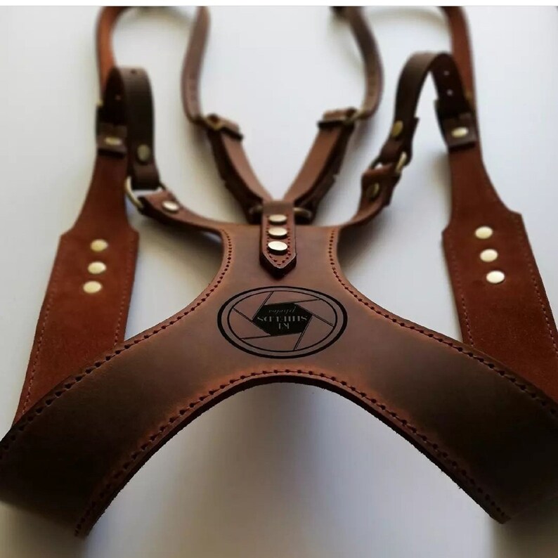 May include: A brown leather camera harness with adjustable straps and brass rivets. The harness features a central chest piece with an oval logo that reads "KL Shields Photography". The design includes multiple connection points for attaching cameras.