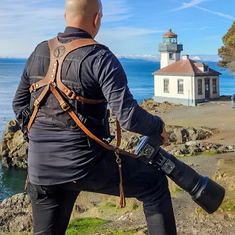 May include: A brown leather camera harness with brass buckles and a logo on the chest. The harness is worn over a black shirt and vest. The photographer is holding a large black camera with a long lens, standing near a lighthouse.