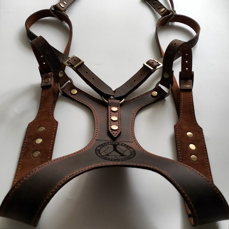 May include: A brown leather harness with adjustable straps and brass-colored hardware. The harness features a chest plate with a circular logo and the text "YORLPHOTOGRAPHY.COM". The design includes multiple straps and rivets.