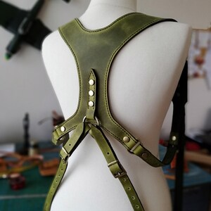 May include: Olive green leather harness with adjustable straps and metal hardware. The harness features a Y-shaped design on the chest and is displayed on a mannequin. The leather has a distressed look, and the stitching is visible.