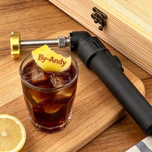 Könnte beinhalten: Ein Cocktail mit Eiswürfeln und einem Zitronenkeil mit dem Text "By-Andy", neben einem schwarzen und goldenen Brenner und einer Holzkiste. Das Getränk befindet sich in einem klaren Glas auf einem Holzschneidebrett.