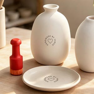 May include: Two matte off-white ceramic vases and a matching ceramic plate, each featuring a circular stamp that reads HANDMADE BY YARUK with a heart icon. A red and orange handheld pottery stamp tool sits to the left of the ceramic plate.