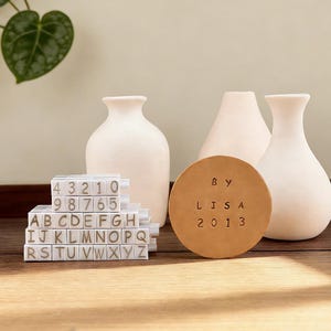 Könnte beinhalten: Ein Stillleben mit weißen Keramikvasen, Buchstaben- und Zahlenblöcken und einem runden Lederuntersetzer. Der Untersetzer ist mit "BY LISA 2013" gestempelt. Die Blöcke zeigen das Alphabet und Zahlen.