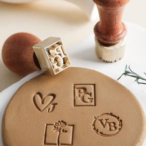 May include: Four different custom stamps on a brown clay surface. The stamps are made of brass and have different designs, including a heart, a monogram, a floral design, and a wreath. The stamps are used to create personalized impressions on clay, paper, or other surfaces.