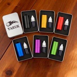 Puede incluir: Una caja metálica plateada con la palabra "YARUK" y un logotipo de pelota de golf. Seis cajas metálicas negras abiertas contienen cada una un tubo de color y una botella pequeña. Los colores incluyen negro, amarillo, rojo, azul, morado y verde.