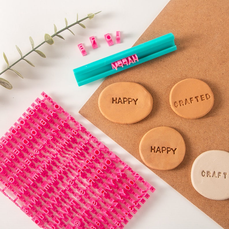 May include: Pink alphabet stamps with a blue and white plastic handle. The stamps are arranged in rows and columns. There are two brown oval-shaped clay pieces with the word "HAPPY" stamped on them. There is a brown oval-shaped clay piece with the word "CRAFTED" stamped on it. There is a white oval-shaped clay piece with the word "CRAFT" stamped on it.