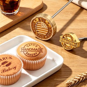 May include: A close-up shot showcases two brass stamps with wooden handles, one featuring an American flag design and the other with text. Two cupcakes with the text "Cato's Handmade Bakery" are in the foreground. A glass of iced tea is in the background.