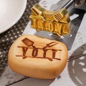 Può includere: Un ferro per marchiare dorato con la parola "YOTT" stampata su un pezzo di pane. Il ferro per marchiare è posizionato sopra il pane, pronto per l'uso. Il pane è marrone chiaro e ha una forma leggermente arrotondata.