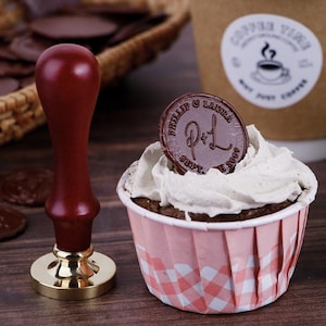 May include: A dark red wax seal stamp with a gold base and a cupcake topped with white frosting and a chocolate seal. The seal has the text "PHILLIP & LAURA" and "P+L SEPT. 2009". A coffee cup is in the background.