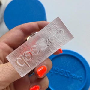 Könnte beinhalten: Ein klarer, rechteckiger Stempel mit dem Wort "Seeb" eingraviert. Der Stempel wird über einem blauen kreisförmigen Objekt gehalten. Der Stempel hat eine strukturierte Oberfläche und wird von einer Person mit orangefarbenem Nagellack gehalten.