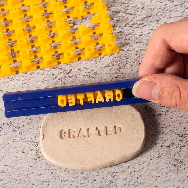May include: A blue and white plastic tool with yellow letters spelling "CRAFTED" is being used to stamp the word "CRAFTED" into a piece of clay.