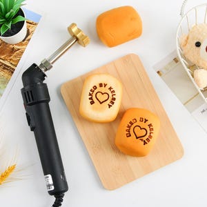 Könnte beinhalten: Ein schwarzes elektrisches Branding-Eisen mit einer Metallspitze und ein Holzbrett mit zwei hellbraunen Brötchen. Die Brötchen sind mit dem Text "Baked by Kelsey" und einem Herzen geprägt.