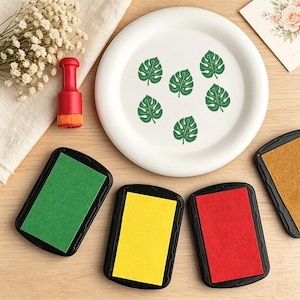 May include: A white ceramic plate featuring six green monstera leaf prints, accompanied by a red handheld stamp tool and four rectangular ink pads in green, yellow, red, and gold, arranged on a light wooden surface with dried flowers.