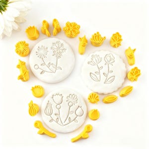 May include: A collection of yellow flower-shaped cookie cutters and three white, round cookies with floral designs. The cutters are in various shapes, including petals, leaves, and complete flower forms. The cookies have detailed floral patterns.
