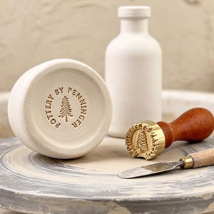 Könnte beinhalten: Ein weißer Keramikgefäßboden mit dem geprägten Text POTTERY BY PENNINGER und einem Tannenbaumsymbol, eine passende weiße Keramikflasche, ein Messingstempel mit Holzgriff und ein Töpferwerkzeug aus Metall mit Holzgriff.