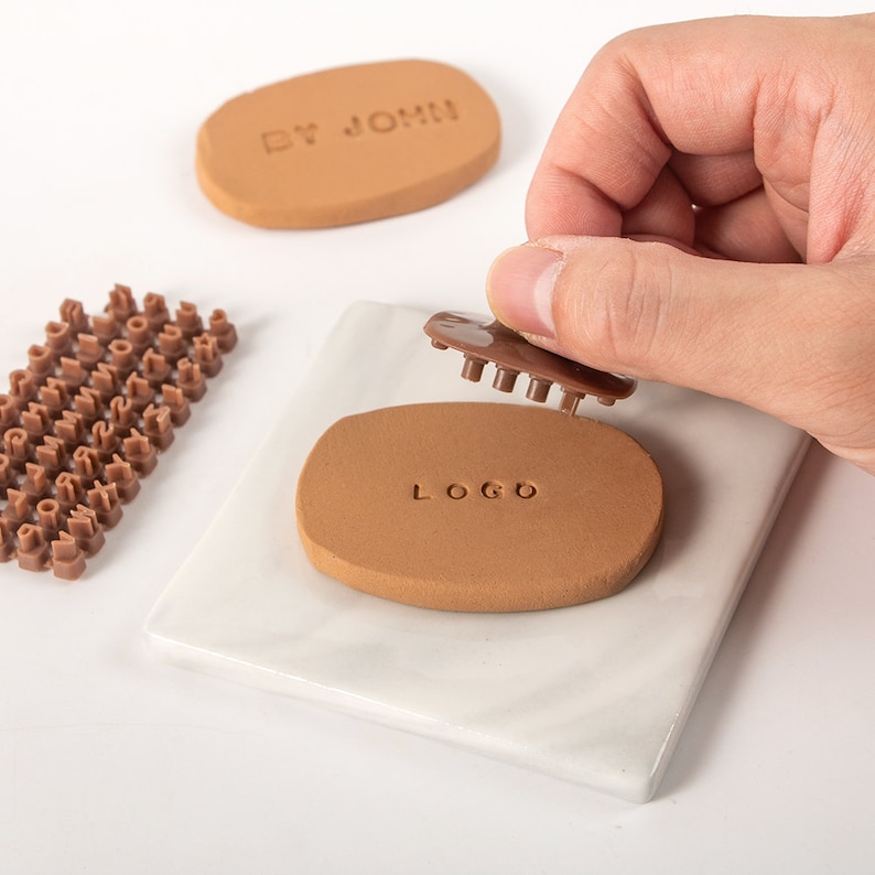 May include: A set of brown plastic alphabet stamps for stamping on clay or other materials. The stamps are arranged in rows on a white surface. A hand is holding a stamp with the word "LOGO" stamped on a brown clay oval.