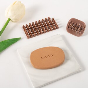 May include: A set of brown alphabet stamps and a brown stamp with the word "LOGO" on it. The stamps are on a white surface with a brown oval-shaped object with the word "LOGO" embossed on it.