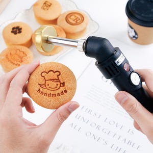 May include: A close-up of a handheld branding iron with a gold-colored head, used to imprint designs onto baked goods. The image shows several round, golden-brown pastries with various stamped designs, including the word "handmade".