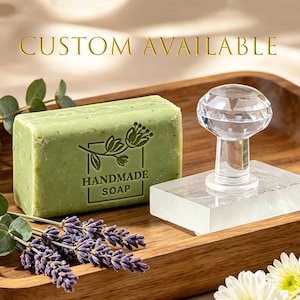 Puede incluir: Una pastilla de jabón rectangular de color verde claro con el texto HANDMADE SOAP y un gráfico floral en relieve en la parte frontal. Junto a ella hay un sello de acrílico transparente con un mango en forma de diamante. El texto CUSTOM AVAILABLE es visible.