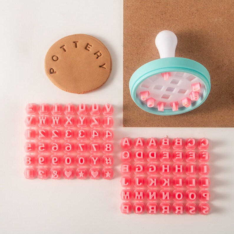 May include: A set of pink alphabet and number stamps for crafting. The stamps are arranged in rows on a white surface. There is a larger blue stamp with a white handle and a grid pattern on the top. The word "POTTERY" is stamped on a brown clay disc.