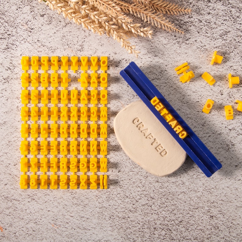 May include: A set of yellow alphabet stamps in a grid, a blue and white tool with the word "CRAFTED" printed on it, and a beige block of clay with the word "CRAFTED" stamped into it. There are also several individual yellow alphabet stamps scattered around the clay.