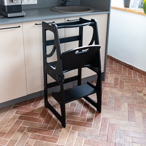 Peut inclure: Un tabouret de cuisine en bois noir avec une marche et une barre de sécurité. Le tabouret est conçu pour aider les enfants à atteindre les plans de travail. L'arrière-plan montre une cuisine avec un évier, des placards et une machine à café.