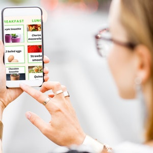 May include: A person is looking at a smartphone screen displaying a meal plan with breakfast and lunch options. The breakfast options include purple smoothie, two boiled eggs, and chocolate smoothie. The lunch options include chorizo mozzarella, strawberry smoothie, and quinoa.