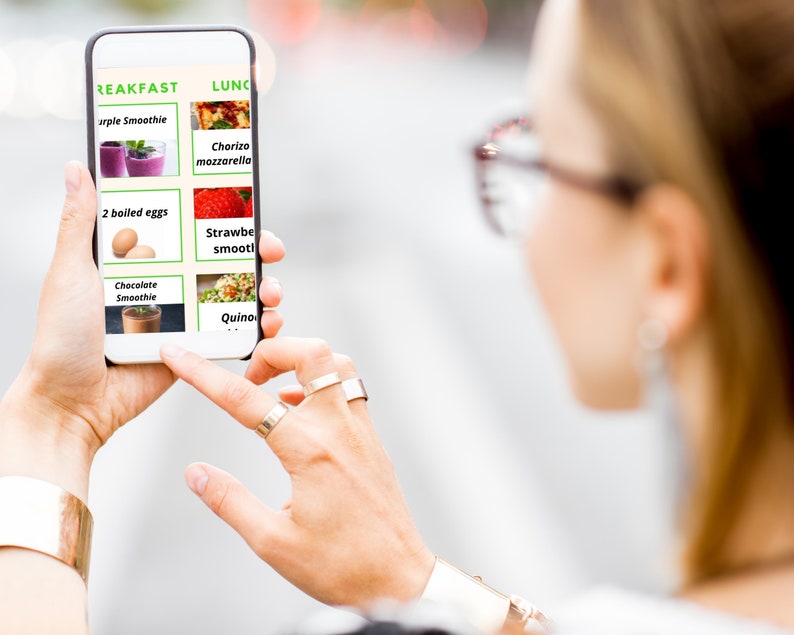 May include: A person is looking at a smartphone screen displaying a meal plan with breakfast and lunch options. The breakfast options include purple smoothie, two boiled eggs, and chocolate smoothie. The lunch options include chorizo mozzarella, strawberry smoothie, and quinoa.