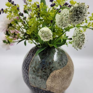 Könnte beinhalten: Eine runde Keramikvase mit einem gesprenkelten teal, dunkelgrauen und beigen Design hält einen Blumenstrauß. Die Blumen umfassen weiße, lila und gelbe Blüten. Die Vase hat eine glänzende Oberfläche und ein einzigartiges, segmentiertes Muster.