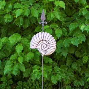 Könnte beinhalten: Ein Gartenstecker mit einer hellen, spiralförmigen Ammoniten-Fossilskulptur, die auf einem dünnen Metallstab montiert ist. Die Spitze des Steckers ist mit einer spitzen, speerartigen Metallverzierung versehen, die einen dekorativen Akzent setzt.