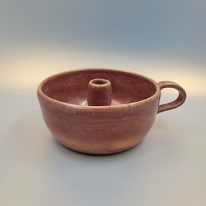 Könnte beinhalten: Ein handgefertigter, rotbrauner Kerzenhalter aus Keramik mit Griff. Der Kerzenhalter hat eine kreisförmige Schalenform mit einer zentralen, zylindrischen Halterung für eine Kerze. Der Griff ist gebogen und an der Seite der Schale befestigt.