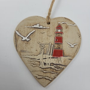 Könnte beinhalten: Ein herzförmiger Keramik-Anhänger mit einem rot-weißen Leuchtturm, einem Steg und Möwen, die über einem blauen Meer fliegen. Der Anhänger hat eine rustikale, handgemalte Optik.