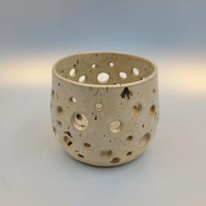 Könnte beinhalten: Ein handgefertigter Keramik-Votivkerzenhalter. Das cremefarbene, zylindrische Gefäß weist zahlreiche kreisförmige Perforationen auf. Dunkelbraune Sprenkel zieren die Oberfläche. Die offene Oberseite lässt das Licht durch die Löcher scheinen.