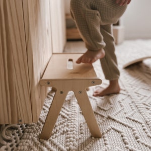 May include: A light wood step stool with a cutout handle on the top. The stool is standing on a white rug with a geometric pattern.