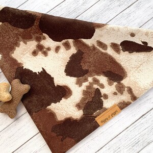 May include: A brown and white cow print dog bandana with a brown leather tag that says "Sips &  Paws".