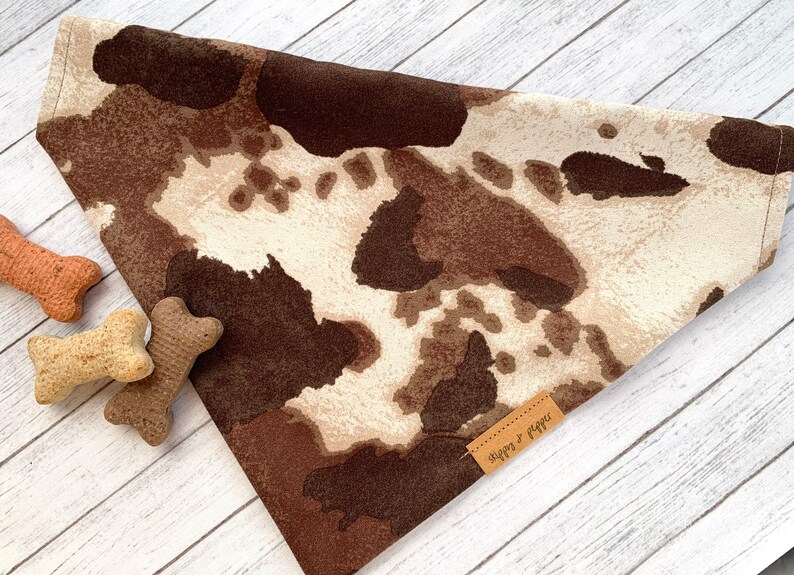 May include: A brown and white cow print dog bandana with a brown leather tag that says "Sips &  Paws".