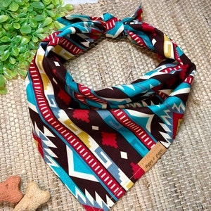 May include: A patterned dog bandana in brown, turquoise, red, and yellow, with a Southwestern design. The bandana is tied at the top. A small leather tag reads "nibbles & kibbles". The bandana's design is geometric.
