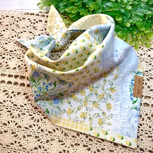 Puede incluir: Un pañuelo para perro de patchwork con estampados florales y de limón en amarillo, azul y blanco. El pañuelo está anudado, mostrando varios diseños de tela. Una pequeña etiqueta marrón está adjunta.