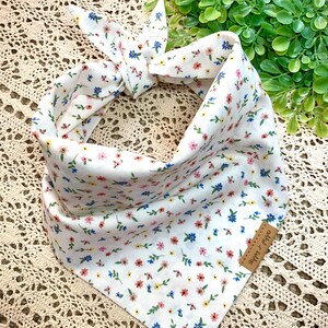 May include: White dog bandana with a floral pattern in pink, blue, and yellow. The bandana is tied in a knot at the top. A small brown leather tag is attached to the bandana.
