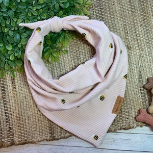 Puede incluir: Un pañuelo para perro rosa con un patrón de abejas. El pañuelo está atado en un nudo y tiene una pequeña etiqueta marrón que dice "Happy Tails".