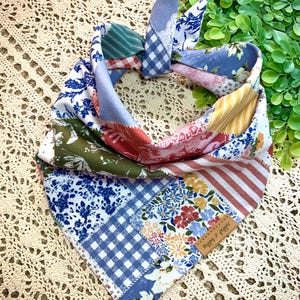May include: A patchwork dog bandana with a variety of floral, striped, and patterned fabrics in shades of blue, red, green, and yellow. The bandana is tied at the top and features a small brown tag with the text "Skippy & Pepper".